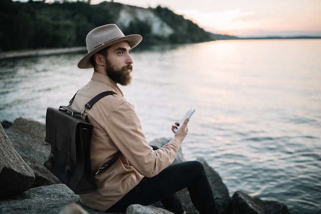 Retrato de jovem homem barbudo segurando o telefone móvel, admirar a natureza e o pôr do sol. Viajante bonito usando chapéu hipster e mochila de couro, sentado na margem do rio. Hora de viajar conceito