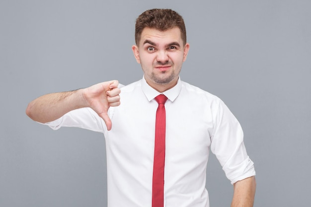 Retrato de jovem insatisfeito de camisa branca e gravata em pé, polegares para baixo e olhando para a câmera com desagrado. interior isolado em fundo cinza.