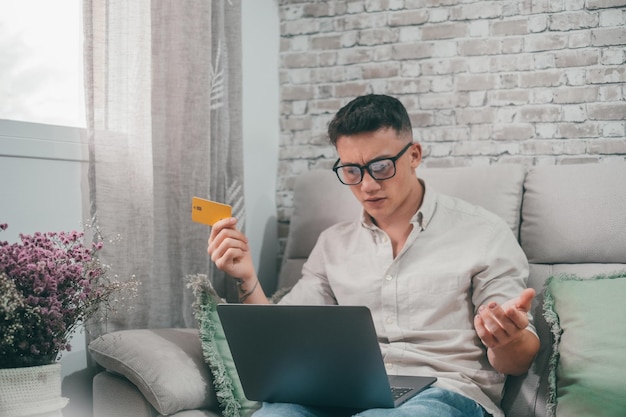 Retrato de jovem irritado e confuso tentando comprar produtos on-line com seu laptop tendo problemas Indivíduo do sexo masculino usando cartão de crédito para fazer compras na internet