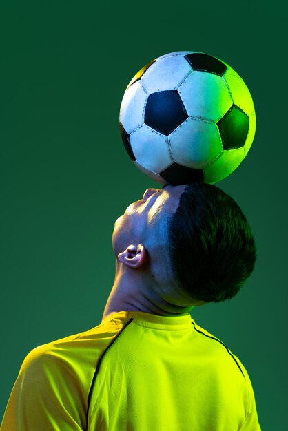 Retrato de jovem jogador de futebol com bola na cabeça posando isolado sobre o fundo verde do estúdio na luz neon