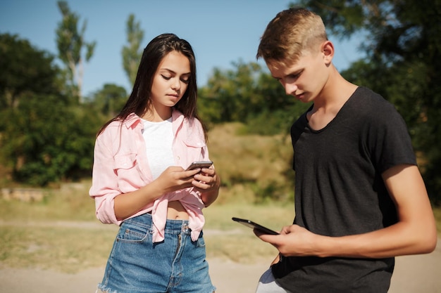 Retrato de jovem lindo menino e menina juntos no parque e olhando pensativamente em celulares