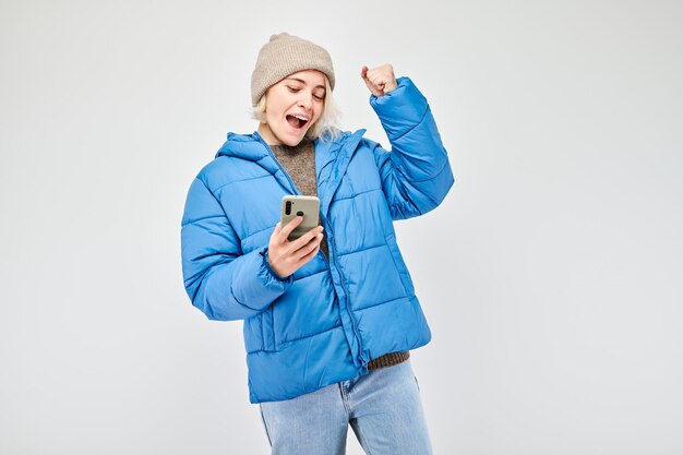 Retrato de jovem loira de jaqueta azul com telefone móvel na mão comemora a vitória ganhando dinheiro no cassino Pessoa com smartphone isolado em fundo branco