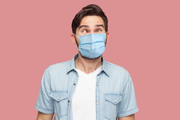 Foto retrato de jovem louco com máscara médica cirúrgica na camisa azul em pé com os olhos vesgos e olhando com cara engraçada de comediante. tiro de estúdio interno, isolado no fundo rosa.