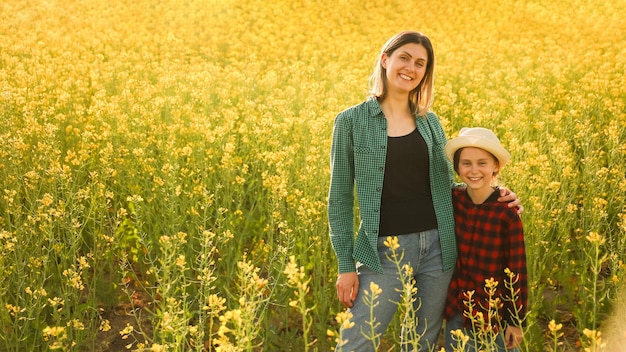 Retrato de jovem mãe mulher de pé em flor amarela colza abraçar filha pequena menina em chapéu