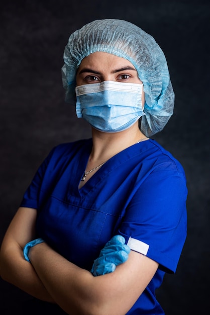 Retrato de jovem médica com máscara cirúrgica protetora vestindo uniforme azul isolado no escuro