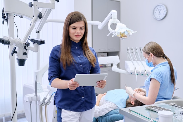 Retrato de jovem médica dentista com tablet digital em consultório odontológico, tratando paciente mulher no fundo da cadeira. Conceito de medicina, odontologia e cuidados de saúde