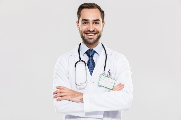 Retrato de jovem médico morena com estetoscópio sorrindo e em pé com os braços cruzados isolados sobre uma parede branca