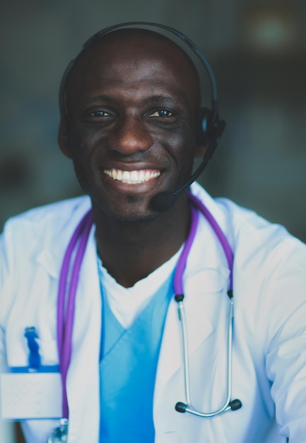 Retrato de jovem médico usando fone de ouvido enquanto estiver usando o computador na mesa na clínica