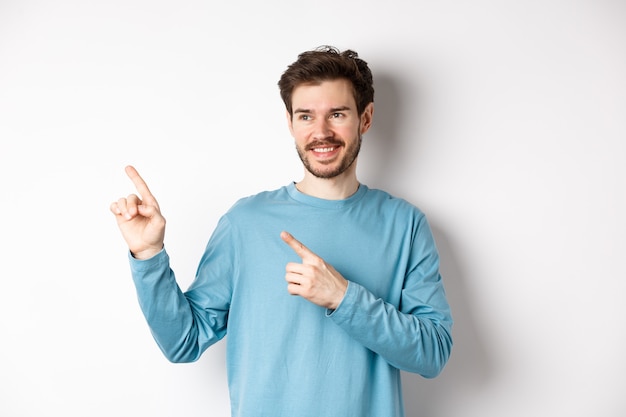 Retrato de jovem moderno em moletom azul apontando os dedos para o canto superior esquerdo e olhando para longe, sorrindo satisfeito por mostrar um anúncio em fundo branco