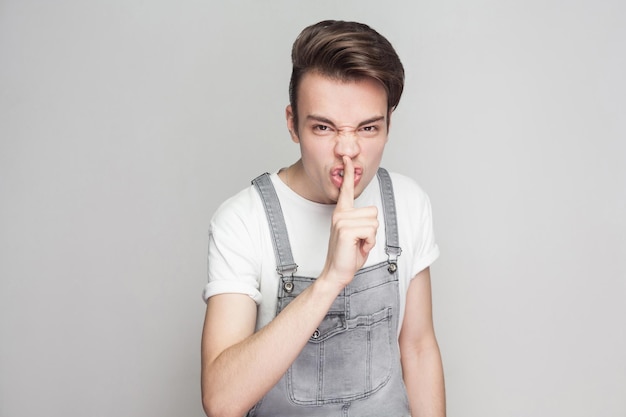 Retrato de jovem morena com raiva em estilo casual com camiseta branca e macacão jeans em pé e olhando para a câmera e mostrando sinal de silêncio. tiro de estúdio interior, isolado em fundo cinza.