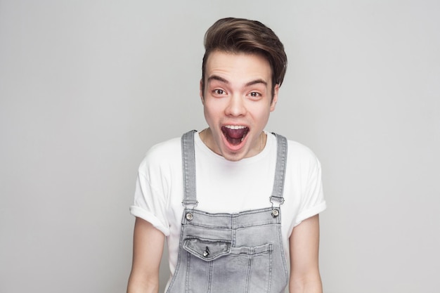 Retrato de jovem morena espantado em estilo casual com t-shirt e macacão jeans em pé e olhando para a câmera com cara de inacreditável surpreso. tiro de estúdio interior, isolado em fundo cinza.