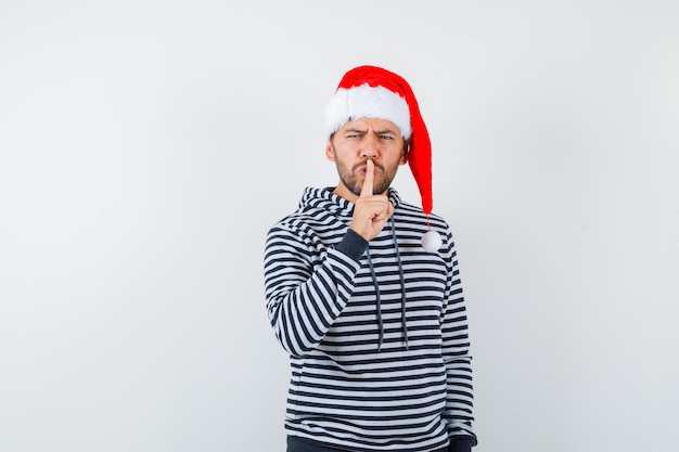 Retrato de jovem mostrando gesto de silêncio com capuz, chapéu de Papai Noel e parecendo irritado