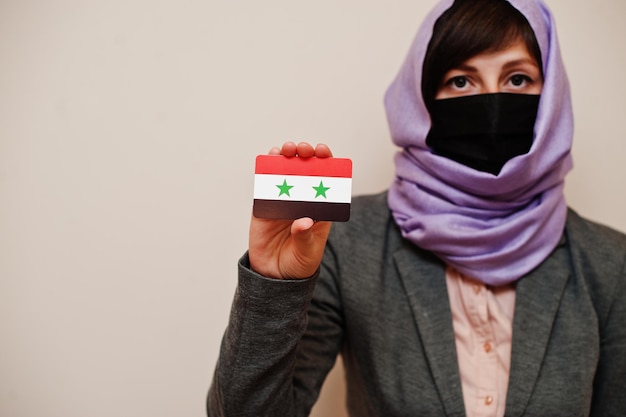Retrato de jovem muçulmana vestindo roupa formal protege máscara facial e lenço de cabeça hijab segura cartão de bandeira da síria contra fundo isolado conceito de país coronavirus