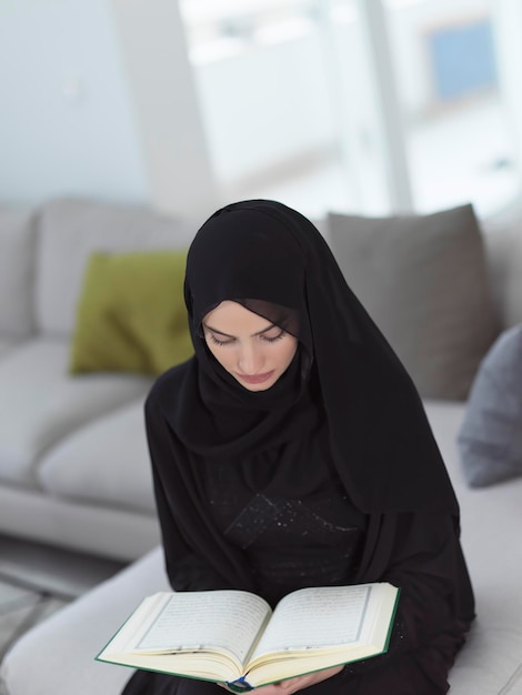 Retrato de jovem muçulmano lendo o Alcorão em casa moderna. Menina árabe recitando o livro sagrado no Islã para adorar a Alá durante o Ramadã.