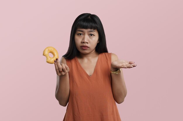 Retrato de jovem mulher asiática olhando rosquinha vitrificada. Maus hábitos de nutrição no conceito de trabalho