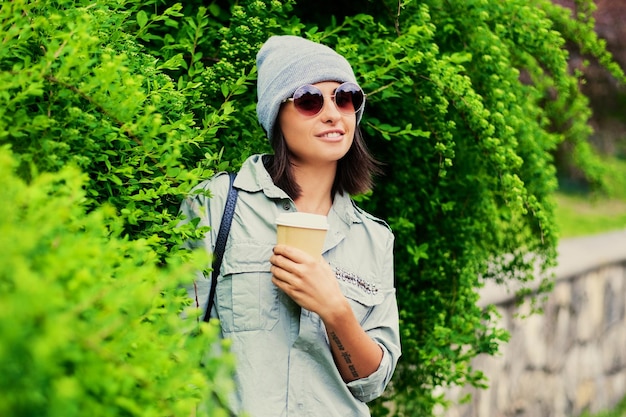 Retrato de jovem mulher atraente em óculos de sol detém xícara de café de papel em um parque verde de verão.