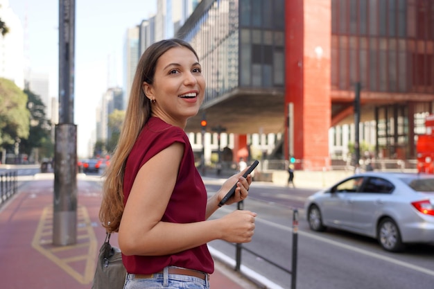 Retrato de jovem mulher de negócios segurando um smartphone olhando para longe na Avenida Paulista, São Paulo, Brasil