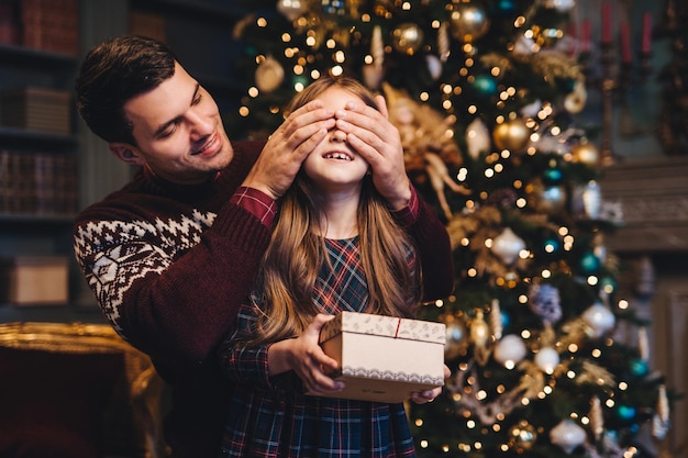 Retrato de jovem pai cobre os olhos de sua filha como vai fazer surpresa para ela dá presente ficar junto perto da árvore de Natal Garota sorridente feliz recebe presente do pai Conceito surpresa