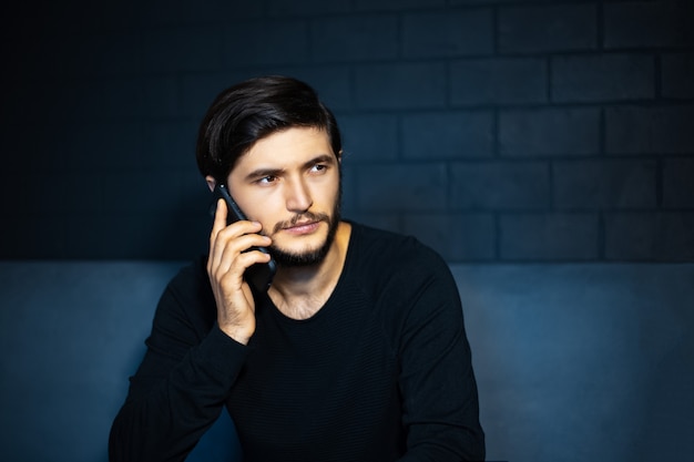 Retrato de jovem pensativo, falando no smartphone, sentado no sofá. parede de tijolos pretos.