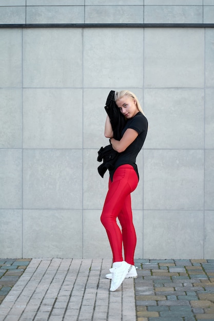 Retrato de jovem positivo desportivo vestindo preto sportswear, leggings vermelhas e tênis brancos na moda. Tiro ao ar livre na parede cinza