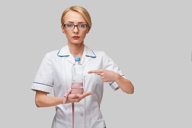 Retrato de jovem profissional de saúde caucasiana segurando uma garrafa de água e fita métrica em pé sobre uma parede cinza claro