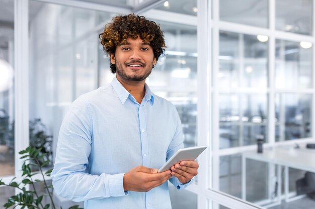 Retrato de jovem programador com computador tablet dentro do escritório homem hispânico com cabelo encaracolado sorrindo