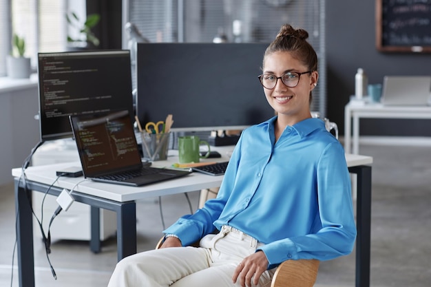 Retrato de jovem programador de óculos sorrindo para a câmera enquanto está sentado no escritório de TI