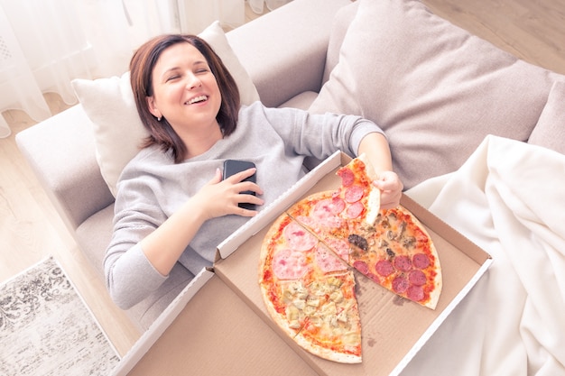 Retrato de jovem rindo deitado no sofá, comendo pizza e segurando o telefone tom laranja