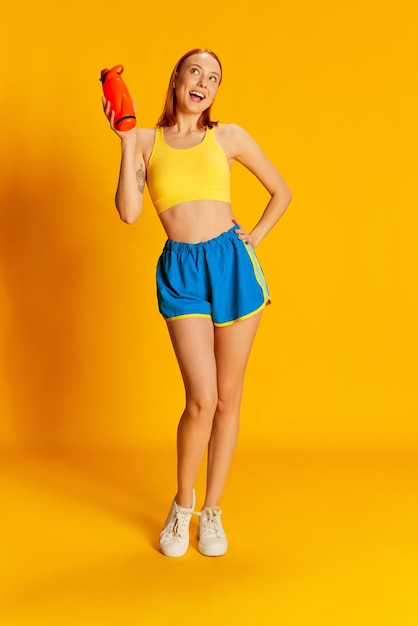 Retrato de jovem ruiva com corpo magro em roupas esportivas posando com garrafa de água sobre fundo amarelo