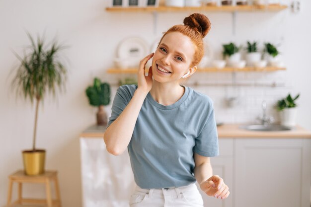 Retrato de jovem ruiva despreocupada feliz ouvindo música em fones de ouvido sem fio enquanto está sentado na mesa olhando para a câmera Mulher sorridente se divertindo enquanto ouve música no escritório em casa