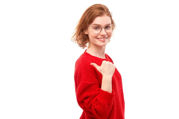 Retrato de jovem ruiva positiva na camisola vermelha de natal sorrindo apontando o dedo para o espaço de cópia vazio para texto ou produto isolado no banner de publicidade de fundo branco