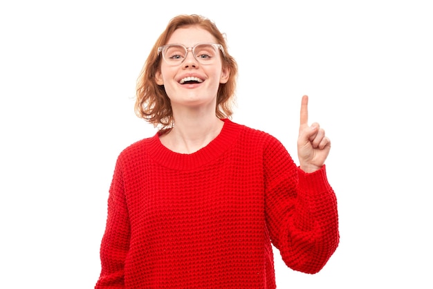 Retrato de jovem ruiva positiva na camisola vermelha de natal sorrindo apontando o dedo para o espaço de cópia vazio para texto ou produto isolado no banner de publicidade de fundo branco