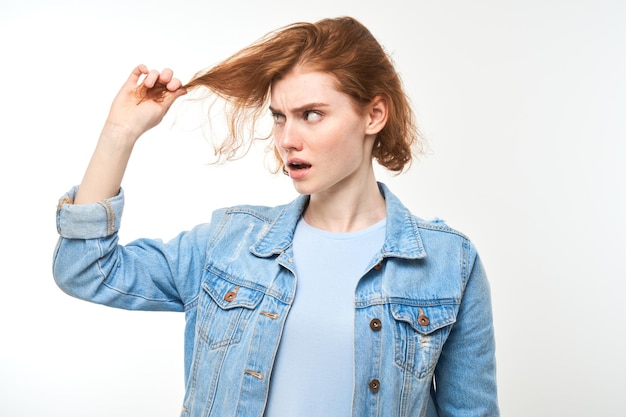 Retrato de jovem ruiva tocando o cabelo com rosto descontente isolado em fundo de estúdio branco conceito de cabelo danificado