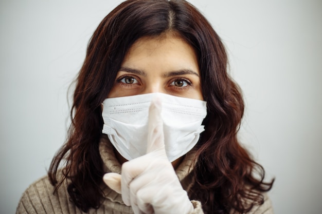 Foto retrato de jovem segura em seu dedo mostrando sinal de silêncio e tem que ficar em casa durante a quarentena devido a pandemia de coronavírus. linda garota usando luvas e máscara médica. epidemia de covid-19.