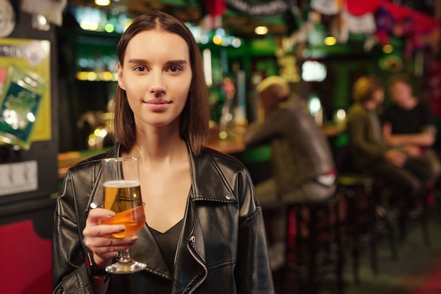 Retrato de jovem segurando um copo de cerveja e olhando para a câmera em pé no bar