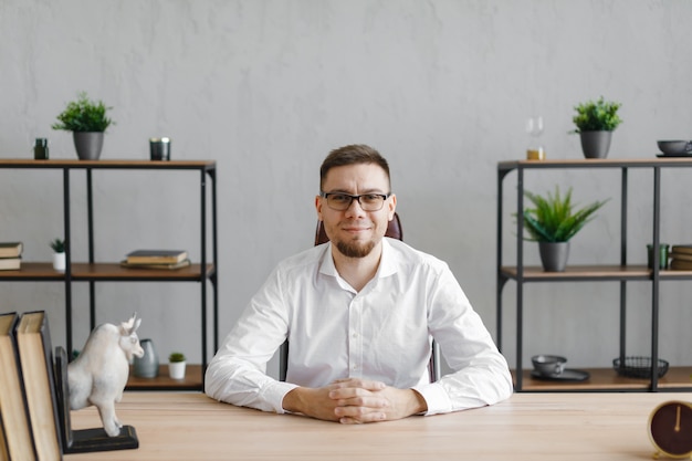 Retrato de jovem sentado em sua mesa no escritório e sorrindo para a câmera enquanto está sentado no local de trabalho