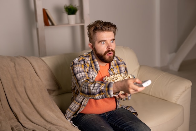 Retrato de jovem sentado no sofá assistindo TV segurando o controle remoto e comendo pipoca