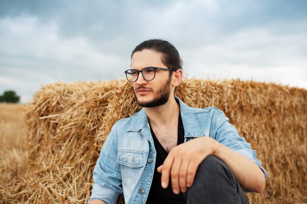 Retrato de jovem sentado perto de montes de feno no fundo das nuvens.