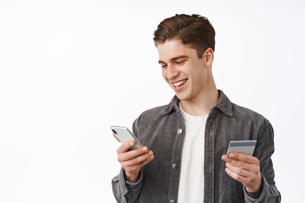 Retrato de jovem sorridente comprando na loja de celular, pagando com smartphone e cartão de crédito, entrega de pedidos no celular, em pé contra um fundo branco