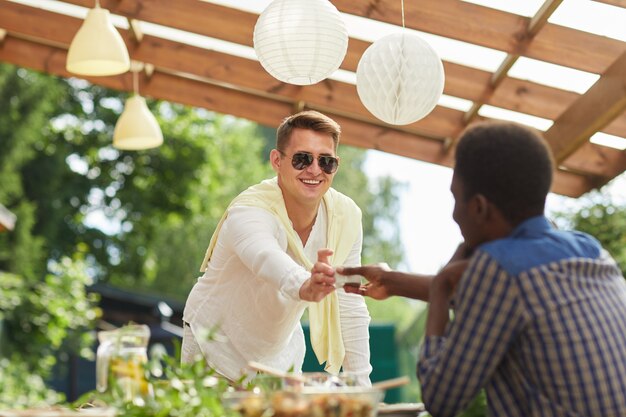 Retrato de jovem sorridente usando óculos escuros, passando o molho para um amigo na mesa enquanto desfruta de um jantar ao ar livre na festa de verão