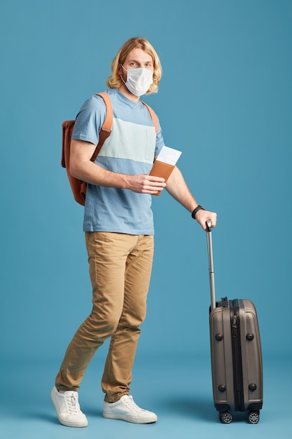 Retrato de jovem turista em pé de máscara protetora com bagagem contra o fundo azul