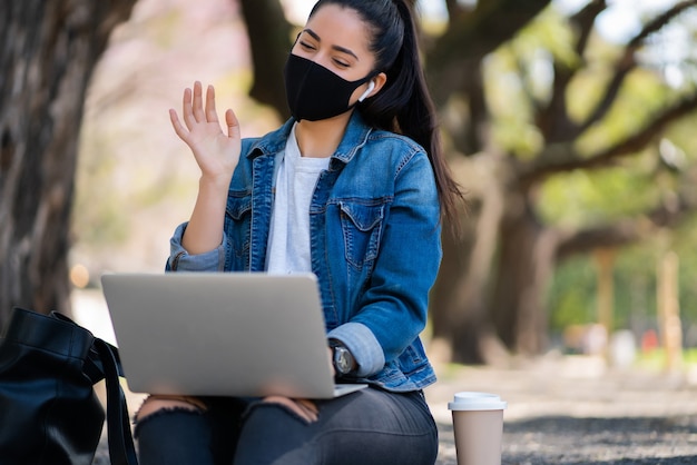 Retrato de jovem usando máscara facial em uma videochamada com laptop enquanto está sentado ao ar livre. conceito urbano. novo conceito de estilo de vida normal.