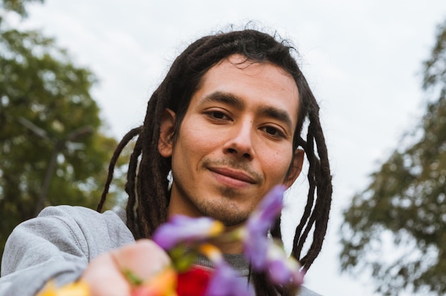 Retrato de jovem venezuelano com dreadlocks sorrindo ao ar livre segurando flores com a mão