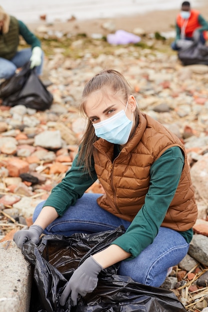 Retrato de jovem voluntário com máscara protetora sentado perto do saco de lixo e olhando para a câmera ao ar livre