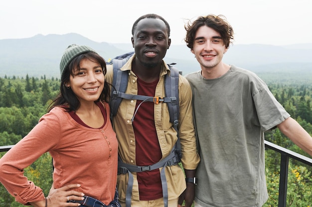 Retrato de jovens amigos multiétnicos positivos em pé na ponte contra a bela paisagem florestal