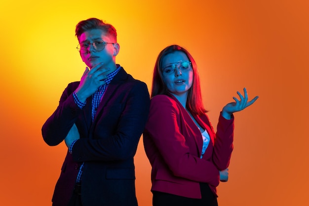 Retrato de jovens empregados homem e mulher posando com expressão facial pensativa isolada sobre fundo laranja em luzes de néon