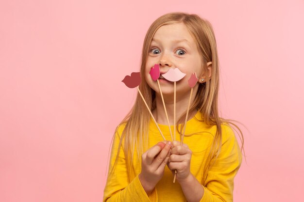 Retrato de linda garotinha surpresa bonitinha olhando com espanto segurando alguns lábios de papel rosa vermelho em palitos escolha de batom cor moda e conceito de cuidados de beleza tiro de estúdio interno isolado