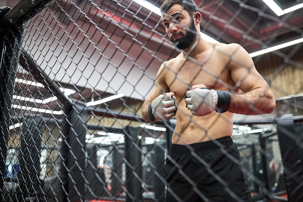 Retrato de lutador de mma pensando em estratégia para a luta, em pé no ringue se preparando para a luta, homem musculoso sem camisa vai praticar luta. atirado através da gaiola