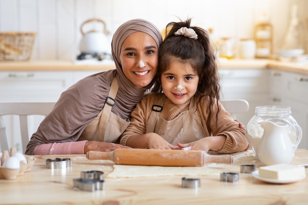 Retrato de mãe de família muçulmana feliz e filhinha assando na cozinha, senhora islâmica e seu filho com farinha no nariz se divertindo enquanto cozinhava pastelaria caseira, aproveitando o tempo juntos