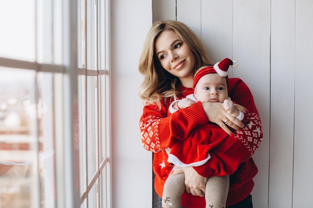 Retrato de mãe e filha em roupas tradicionais de natal em sala de madeira clara perto do espaço de cópia da janela